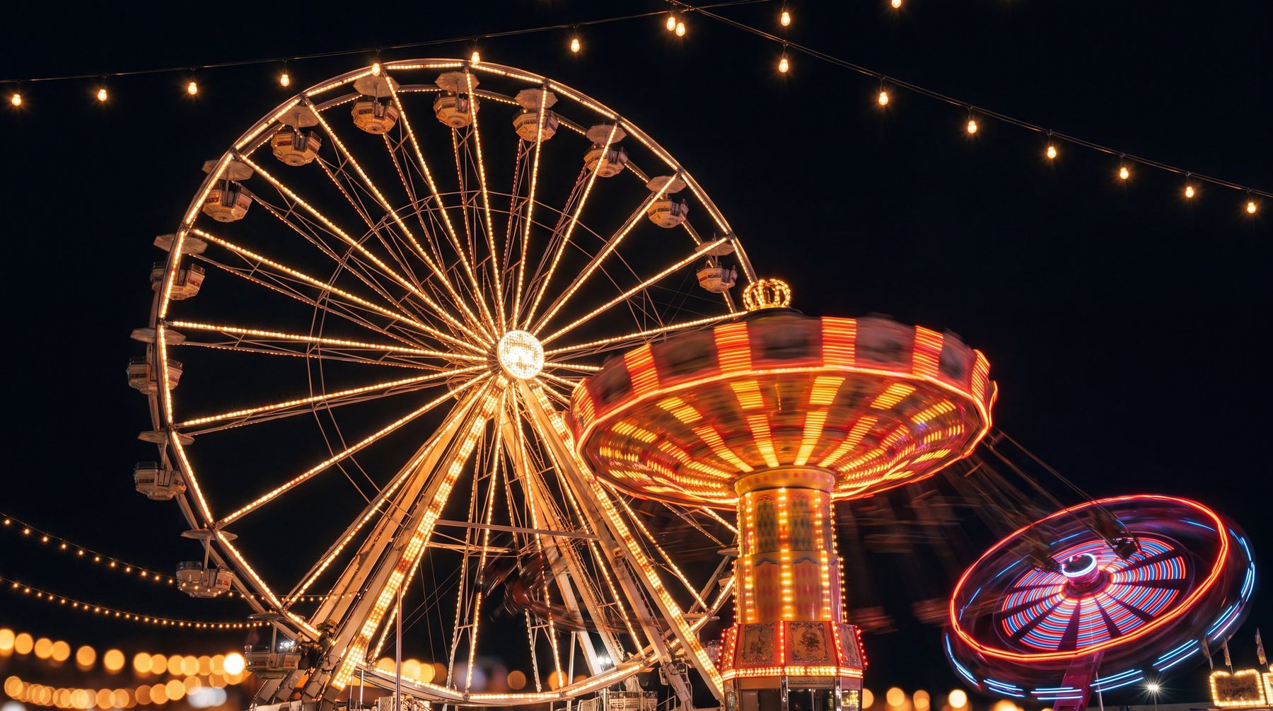 A county fair at night: a ferris wheel and a swing carousel lit up in amber bulbs against a black sky, with strings of lights overhead.
